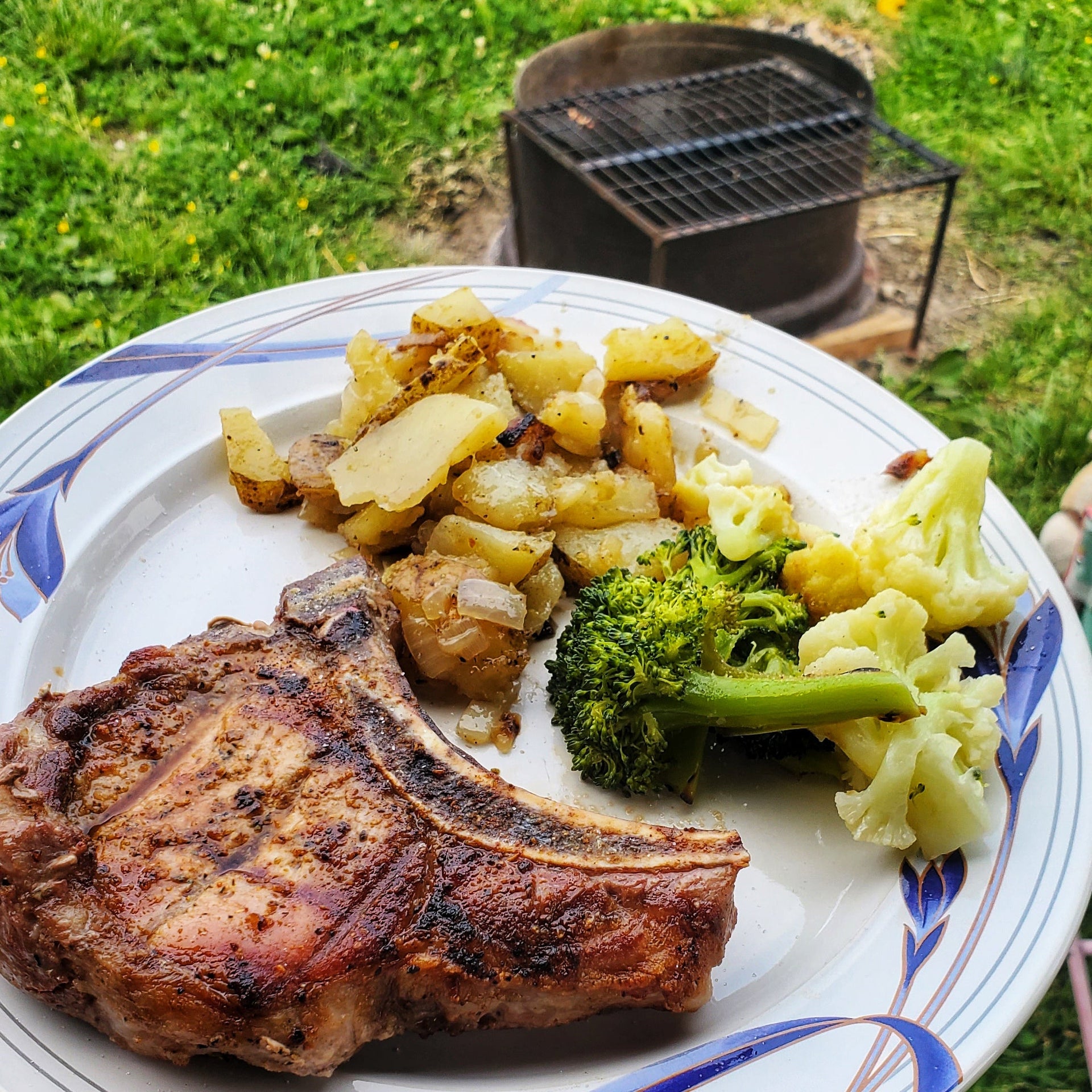 A cooked cjw regenerative bone in pork chop with roasted potatoes and broccoli with a fire pit in the background